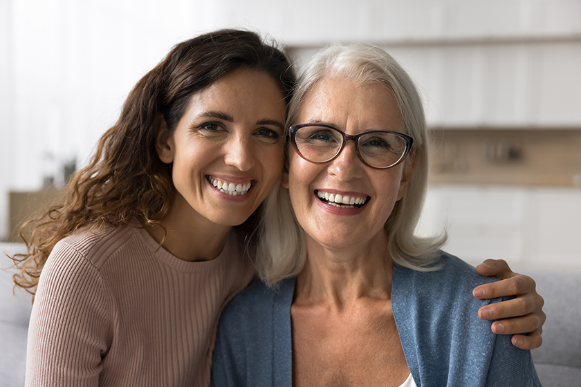 Cheerful beautiful adult daughter woman hugging happy blonde senior mom, holding shoulder with support, care, looking at camera with perfect toothy smile, laughing. Elderly mother headshot portrait 