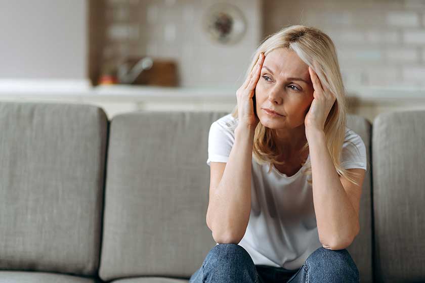 Sad caucasian senior woman wearing casual clothes sits on couch at home alone feels unhappy because of headache, stress, illness or bad news, she needs rest and sleep